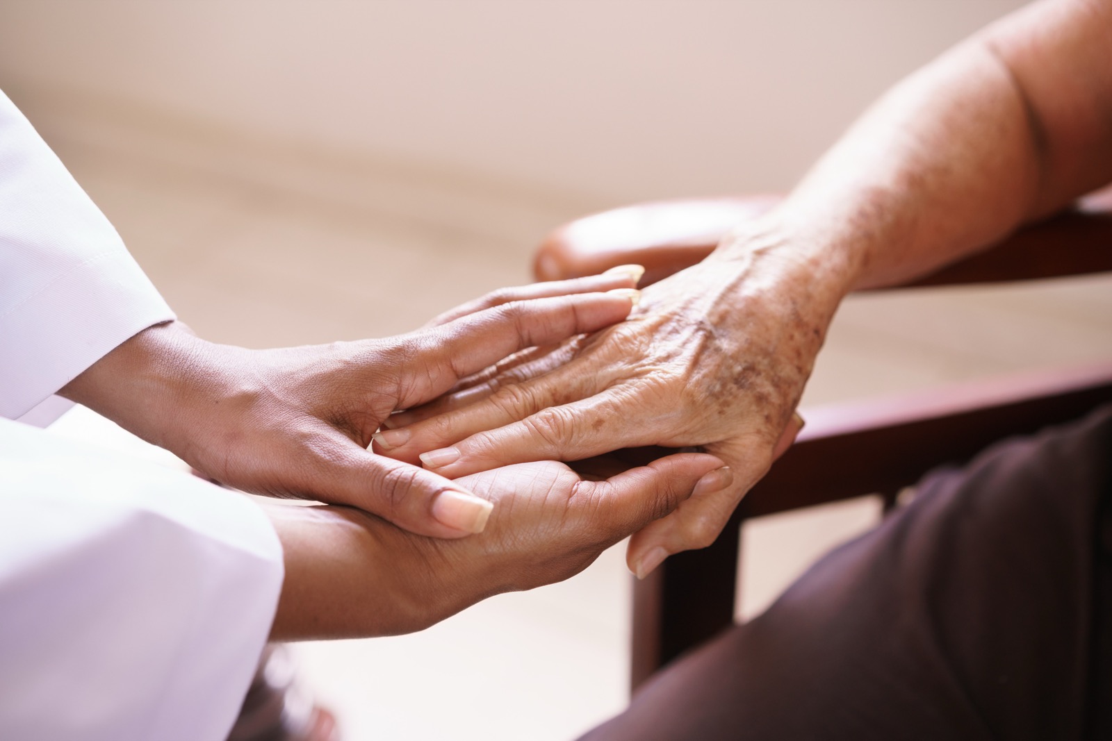 A caregiver's hands cradle an elderly person's hand at home, a quiet gesture of continuity and trust.
